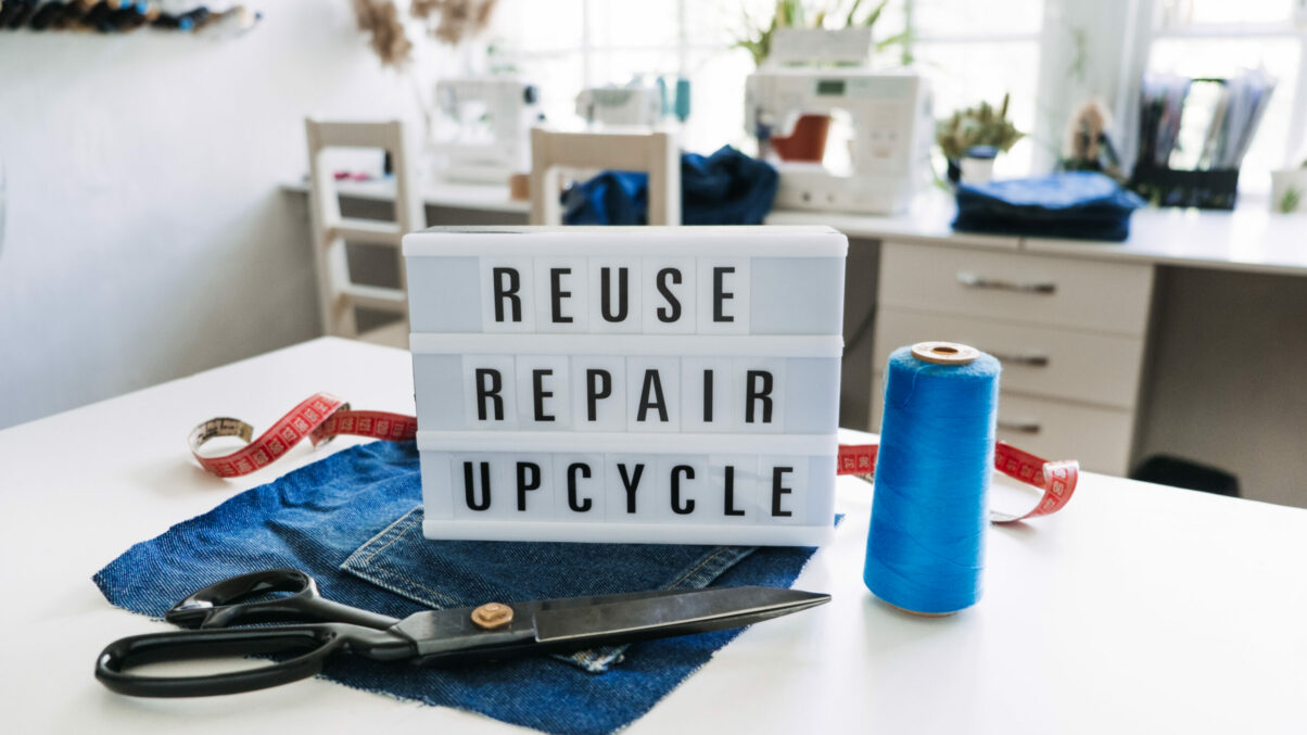 Wiederverwenden, reparieren, Upcycling-Text auf Leuchttafel auf Nähmaschinenhintergrund. Stapel alter Jeans, Jeanskleidung, Scheren, Garn und Nähwerkzeuge im Nähstudio. Denim-Upcycling-Ideen mit alten Jeans.