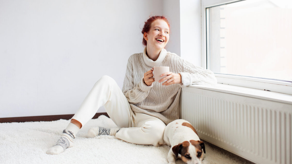 Junge Frau sitzt mit Hund und Kaffeetasse beim Heizkörper vor dem Fenster.